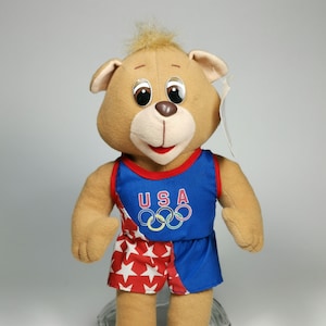 Può includere: Un orsacchiotto di peluche marrone che indossa un completo sportivo blu e rosso con stelle bianche. L'abito ha la scritta "USA" e gli anelli olimpici sul davanti.