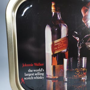 Op de afbeelding: Een metalen dienblad met een gouden rand en een zwarte achtergrond waarop een fles Johnnie Walker Red Label Scotch Whisky is afgebeeld. Het dienblad heeft de tekst "Johnnie Walker, 's werelds best verkopende Scotch whisky" en een rood stick figure dat loopt.