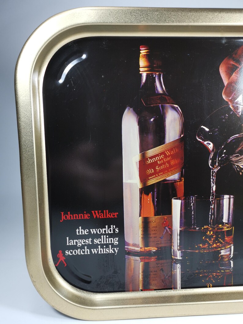 Op de afbeelding: Een metalen dienblad met een gouden rand en een zwarte achtergrond waarop een fles Johnnie Walker Red Label Scotch Whisky is afgebeeld. Het dienblad heeft de tekst "Johnnie Walker, 's werelds best verkopende Scotch whisky" en een rood stick figure dat loopt.