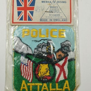 May include: A blue and white embroidered patch with the word "POLICE" in yellow lettering above a green field with three flags. The flags are the American flag, a flag with a yellow circle and red and white stripes, and a flag with a red cross on a white background. The words "ATTALLA" are in yellow lettering below the flags.