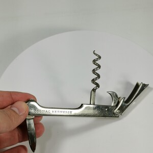 May include: Silver-toned multi-tool corkscrew with a built-in bottle opener and a small knife. The tool is engraved with the words "Cognac Hennessy".