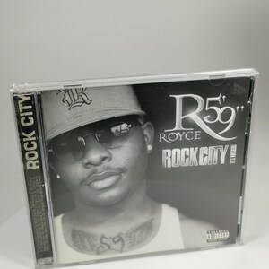 Può includere: Una copertina di CD in bianco e nero con un rapper che indossa un cappello da baseball e occhiali da sole. Il titolo dell'album è "Rock City" di Royce da 5'9".