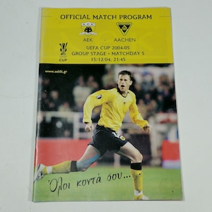 Op de afbeelding: Een geel en zwart voetbalprogramma voor de UEFA Cup-wedstrijd tussen AEK Athene en Alemannia Aachen. Het programma bevat een foto van een voetballer in een geel shirt dat op het veld rent. De tekst op het programma luidt "OFFICIAL MATCH PROGRAM" en "UEFA CUP 2004-05 GROUP STAGE MATCHDAY 5 15/12/04, 21:45".