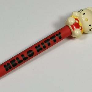 May include: A red pencil with the words "HELLO KITTY" in black. The pencil has a white eraser and a Hello Kitty figure at the top, wearing a red dress and a yellow bow.