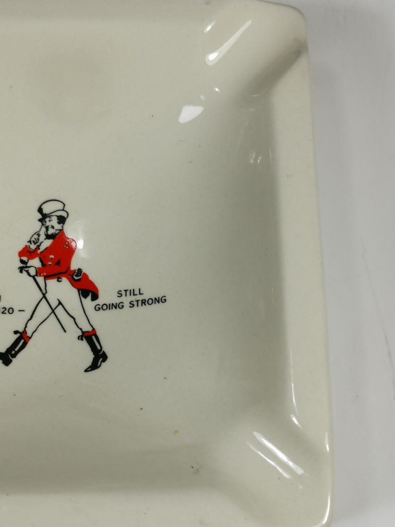 Puede incluir: Cenicero de cer&aacute;mica blanca con una ilustraci&oacute;n en blanco y negro de un hombre con un abrigo rojo y un sombrero de copa. El texto "STILL GOING STRONG" est&aacute; impreso debajo de la ilustraci&oacute;n.