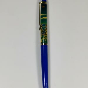 Op de afbeelding: Een blauwe en gouden pen met een groen en gouden ontwerp met een cartoonfiguur. De pen heeft een intrekbare punt en een clip.