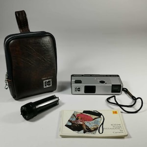 May include: A vintage Kodak Instamatic 10 camera set. The set includes a silver camera, a brown leather case, a black flash attachment, and a booklet. The camera has a black wrist strap. The case has a zipper closure and a handle.