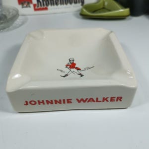Puede incluir: Cenicero de cer&aacute;mica blanca con un logotipo de Johnnie Walker rojo y negro y un gr&aacute;fico de un hombre caminando. El logotipo dice "JOHNNIE WALKER".