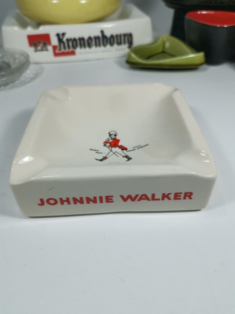 Puede incluir: Cenicero de cer&aacute;mica blanca con un logotipo de Johnnie Walker rojo y negro y un gr&aacute;fico de un hombre caminando. El logotipo dice "JOHNNIE WALKER".