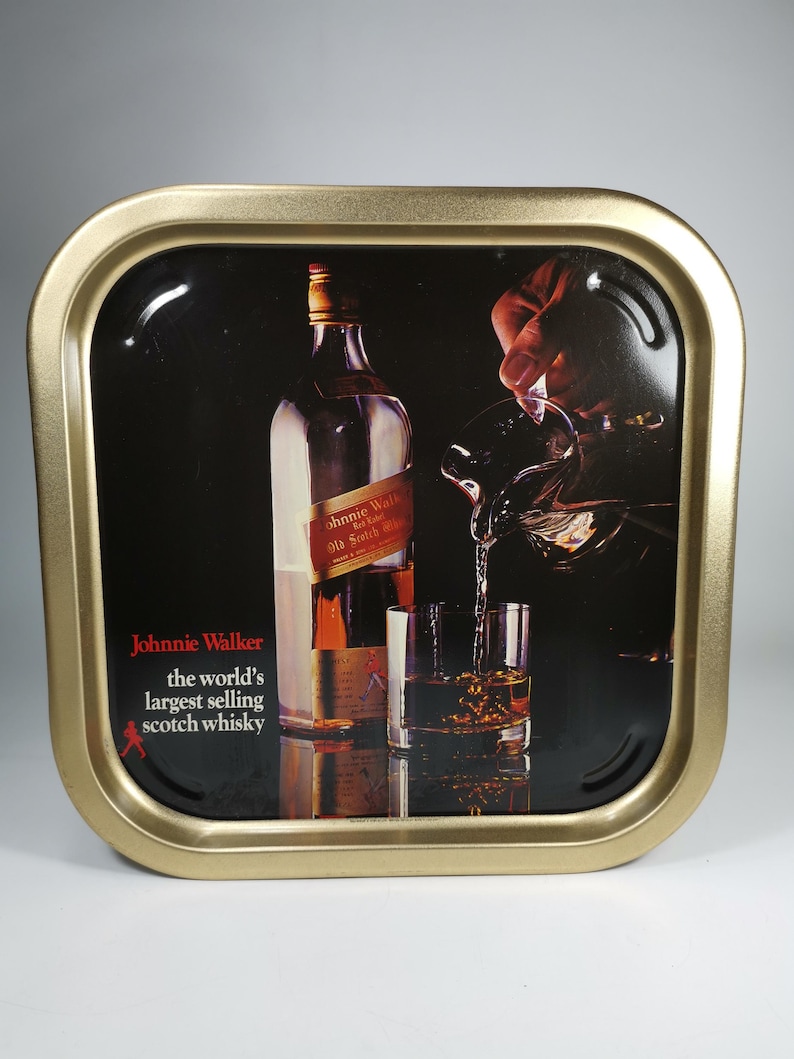 Op de afbeelding: Een gouden metalen dienblad met een zwarte achtergrond waarop een fles Johnnie Walker Old Scotch Whisky in een glas wordt gegoten. De tekst "Johnnie Walker, 's werelds best verkopende Scotch whisky" staat op het dienblad gedrukt.