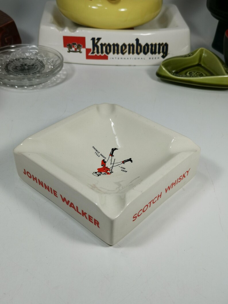 Puede incluir: Cenicero de cer&aacute;mica blanca con un logotipo rojo de Johnnie Walker y las palabras "Scotch Whisky". Una imagen de dibujos animados de un hombre con falda escocesa roja est&aacute; en el centro del cenicero.