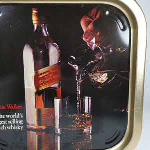 Op de afbeelding: Een gouden metalen dienblad met een zwarte achtergrond waarop een fles Johnnie Walker Red Label Scotch Whisky in een glas wordt gegoten. De tekst "Johnnie Walker Red Label Old Scotch Whisky" is zichtbaar op de fles. De tekst "Johnnie Walker" is zichtbaar in de linkerbenedenhoek van het dienblad. De tekst "The world's best selling Scotch whisky" is zichtbaar onder de tekst Johnnie Walker.