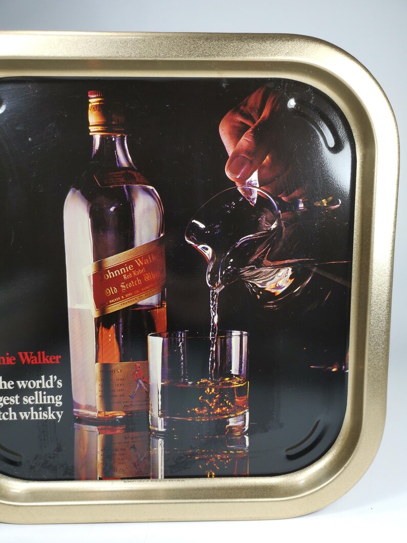 Op de afbeelding: Een gouden metalen dienblad met een zwarte achtergrond waarop een fles Johnnie Walker Red Label Scotch Whisky in een glas wordt gegoten. De tekst "Johnnie Walker Red Label Old Scotch Whisky" is zichtbaar op de fles. De tekst "Johnnie Walker" is zichtbaar in de linkerbenedenhoek van het dienblad. De tekst "The world's best selling Scotch whisky" is zichtbaar onder de tekst Johnnie Walker.