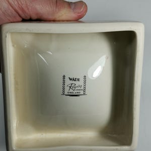 Puede incluir: Un plato cuadrado de cer&aacute;mica blanca con un logotipo en blanco y negro que dice "Wade Reglar London England" dentro de una corona de hojas.