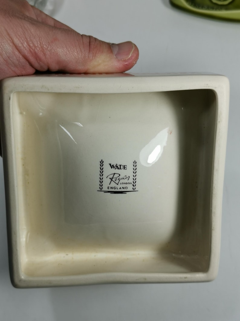 Puede incluir: Un plato cuadrado de cer&aacute;mica blanca con un logotipo en blanco y negro que dice "Wade Reglar London England" dentro de una corona de hojas.