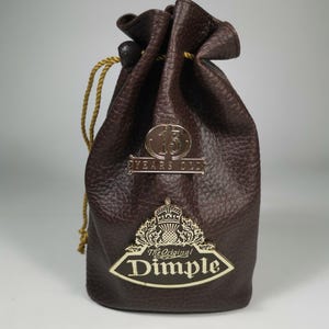Könnte beinhalten: Eine braune Ledertasche mit einem goldenen Kordelzug und einem goldenen Metallemblem, auf dem "The Original Dimple" und "15 Years Old" steht.
