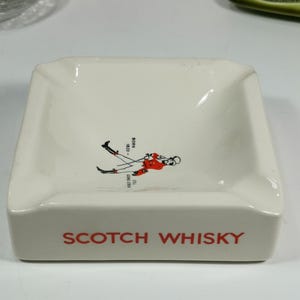 Puede incluir: Cenicero de cer&aacute;mica blanca con una ilustraci&oacute;n roja y negra de una mujer con falda escocesa sosteniendo un palo de golf. El texto "SCOTCH WHISKY" est&aacute; impreso en rojo en la parte delantera del cenicero.