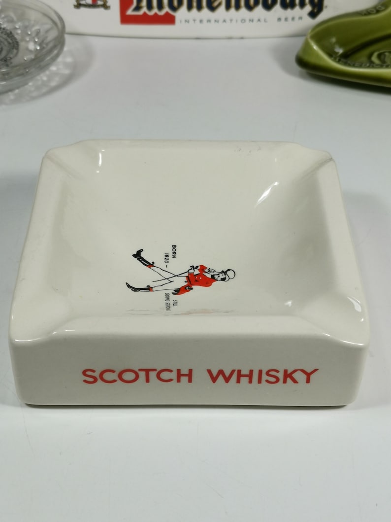 Puede incluir: Cenicero de cer&aacute;mica blanca con una ilustraci&oacute;n roja y negra de una mujer con falda escocesa sosteniendo un palo de golf. El texto "SCOTCH WHISKY" est&aacute; impreso en rojo en la parte delantera del cenicero.