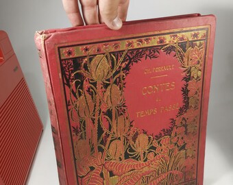 May include: A vintage book with a red cover and gold floral illustrations. The title "Contes du Temps Passé" is visible. The book is held by a hand, suggesting it's being presented or examined. The book's design is ornate and classic.