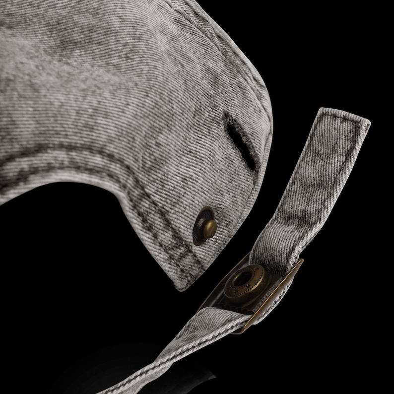 Op de afbeelding: Close-up van een grijze denim baseballpet met een messingknop en een verstelbare band. De band is gemaakt van hetzelfde denimmateriaal als de pet.