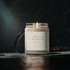 May include: A beige soy wax candle with a clear glass jar and a label that reads "Smells Like Richard Gere" and "Soy Wax Candle". The candle is lit and the flame is visible.