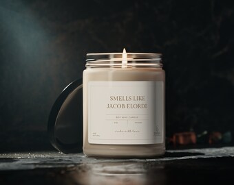 May include: A soy wax candle in a clear glass jar with a beige label that reads "Smells Like Jacob Elordi" and "Soy Wax Candle". The candle is lit and the flame is visible.