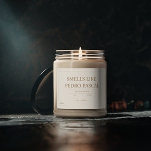 May include: A beige soy wax candle with a clear glass jar and a label that reads "Smells Like Pedro Pascal" in gold lettering. The candle is lit and the flame is visible.