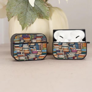 May include: Two black AirPod cases with a colorful book stack design. One case is closed, and the other is open, revealing white earbuds. A white sunflower and pumpkin are in the background.