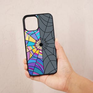May include: A black phone case with a two-part design. One side features a colorful stained-glass pattern, while the other displays a gray spiderweb design. The case is held in a hand.