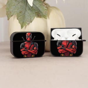 May include: A black and red wireless earphone case with a graphic of Deadpool, a superhero character, with crossed arms and a black background.