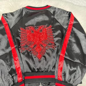 Könnte beinhalten: Schwarze Satin-Bomberjacke mit roten Besätzen und einem großen roten, mit Pailletten besetzten albanischen Adler auf der Rückseite.