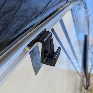 May include: A black plastic hook attached to a silver metal track on a white surface. The hook is designed to hold items such as a towel or a bag.