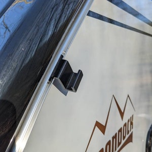 May include: A black, angular RV awning support bracket attached to the side of a white RV. The RV has a brown mountain graphic and the word "BOONDOCK" in brown lettering.
