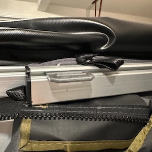 May include: Close-up of a gray zipper on a black and green tent. The zipper is secured with a metal slider and has a brown fabric trim.