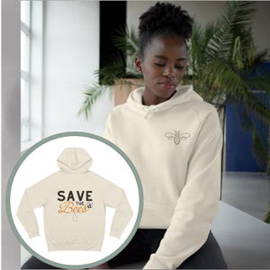 Könnte beinhalten: Ein cremefarbener Hoodie mit einem Bienendesign auf der Vorderseite und dem Text "Save the Bees" auf der Rückseite.
