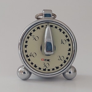 May include: A vintage chrome stopwatch with a cream-colored face and black markings. The stopwatch has a large, central hand and a small Swiss flag logo. It is supported by three spherical feet and has a ring at the top.