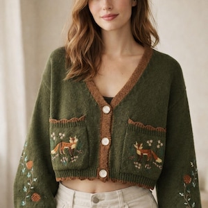 Może przedstawiać: Oliwkowy kardigan z dekoltem w serek i brązowym wykończeniem. Sweter posiada haftowane lisy i kwiatowe detale na kieszeniach i rękawach. Ma białe guziki i jest zestawiony z kremowymi spodniami.