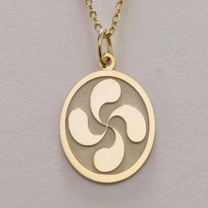 May include: A gold-colored pendant necklace featuring a circular pendant with a stylized four-part design. The pendant hangs from a delicate gold chain. The design is a symbol of Basque culture.