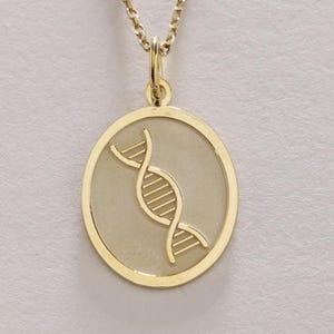 Può includere: Una collana color oro con un ciondolo raffigurante una doppia elica del DNA. Il ciondolo è circolare, con uno sfondo strutturato e una catena sottile. La collana è mostrata su uno sfondo neutro.