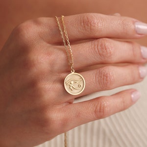 Op de afbeelding: Een gouden ketting met een ronde hanger met een gegraveerd oog van Horus-symbool.