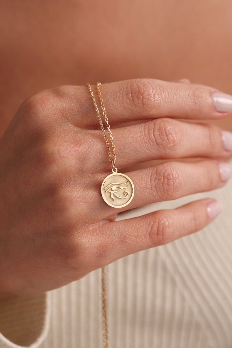 Op de afbeelding: Een gouden ketting met een ronde hanger met een gegraveerd oog van Horus-symbool.