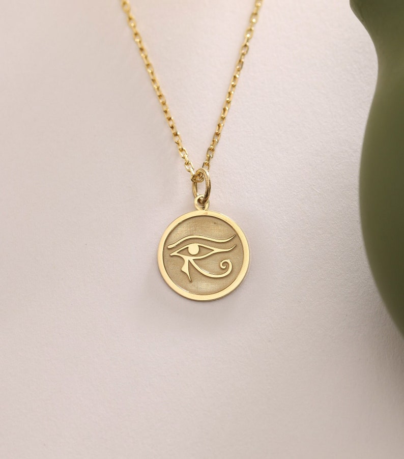 Op de afbeelding: Een gouden ketting met een ronde hanger met een gegraveerd beeld van het oog van Horus.