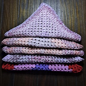 May include: A stack of five crocheted triangular blankets in shades of purple, pink, and red. The blankets are folded in a triangle shape and are arranged in a stack.