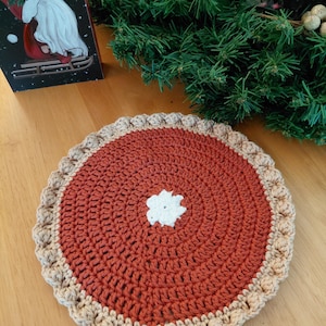 May include: A handmade, crocheted trivet resembling a pie. The center is a warm, reddish-brown color, with a white flower-shaped accent in the middle. The crust is a light tan color with a scalloped edge. The trivet is on a wooden surface.