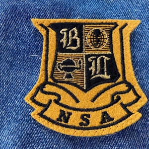 May include: A gold and black embroidered patch with a crest featuring a globe, a cup, and the letters "B.O.L." and "I.L."  The patch has a banner with the letters "NSA" in black.