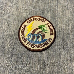 May include: A circular embroidered patch with the words "GULFCOAST COUNCIL HURRICANE PREPAREDNESS". The patch depicts a yellow lightning bolt, palm trees, and blue waves. It has a brown border and is set against a blue denim backdrop.