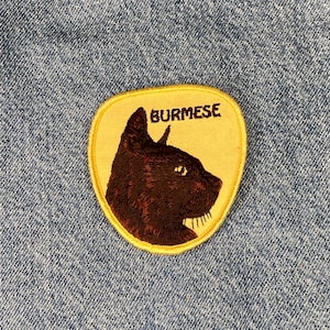 May include: A yellow and brown embroidered patch with a cat's head and the word "BURMESE" in brown lettering.
