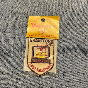 May include: A shield-shaped embroidered patch in a clear plastic sleeve with a yellow header card labelled Voyageur World Wide Collectors Series. The patch features the text Brantford Ont. 1875 First Telephone with a graphic of a vintage telephone.