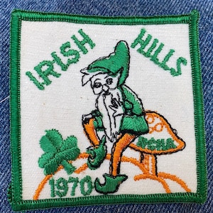 May include: A green and white embroidered patch with a cartoon leprechaun sitting on a mushroom. The patch says "Irish Hills 1970" in green embroidery.