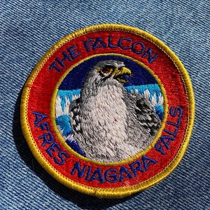 May include: A red, white, and blue embroidered patch with a yellow border. The patch features a white falcon with black markings and the text "The Falcon After Niagara Falls".